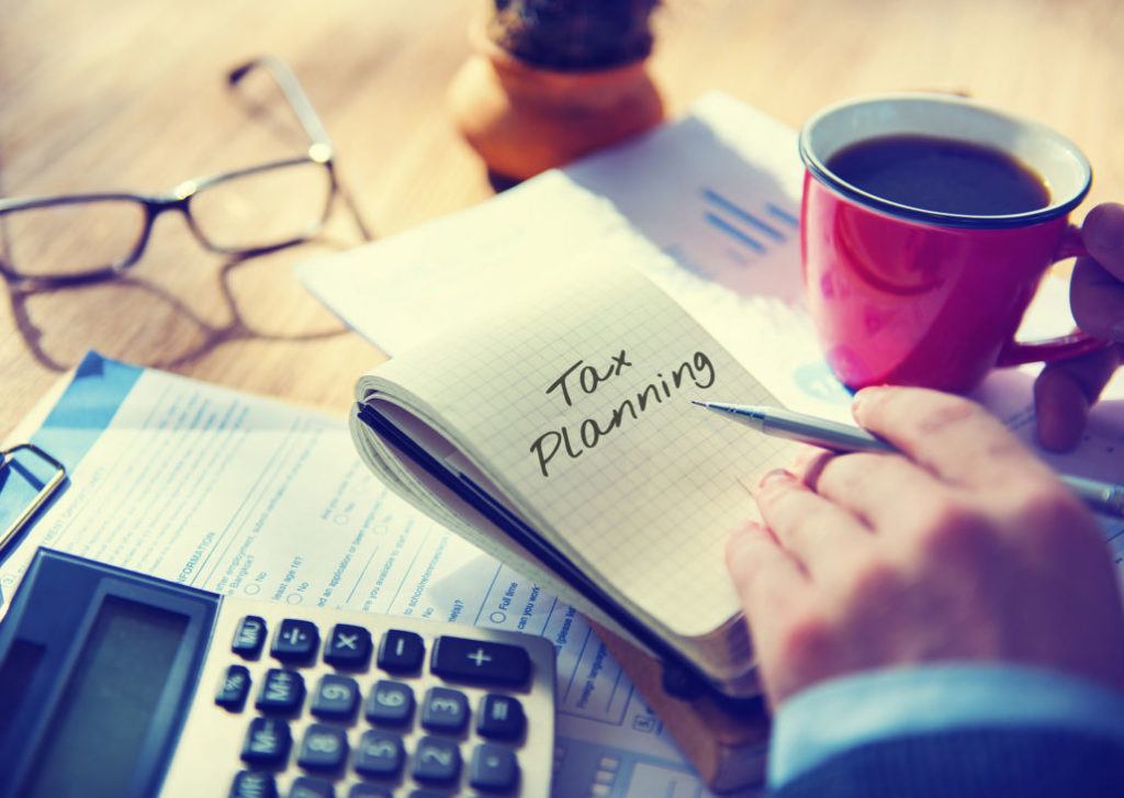 A hand holding a pen, leaning on a table with a calculator, glasses and paperwork on it, laid around a notebook that has "Tax Planning" written on it